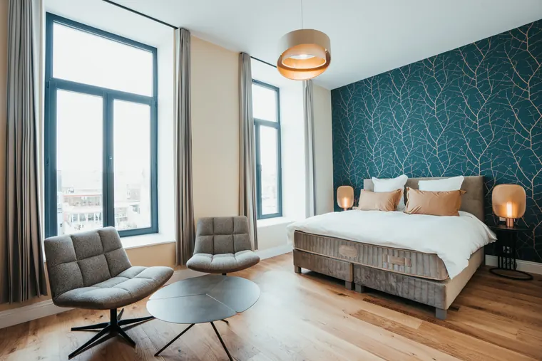 Chambre Prestige — mur bleu canard à branches cuivrées, lit capitonné entre deux hautes fenêtres et coin salon